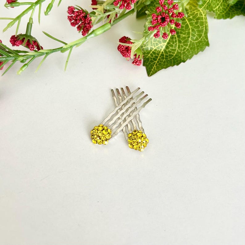 May include: Two silver-toned hair pins featuring circular decorative tops encrusted with small, bright yellow rhinestones. The pins have wavy metal prongs designed for securing hair, and the yellow stones create a textured, sparkling appearance.