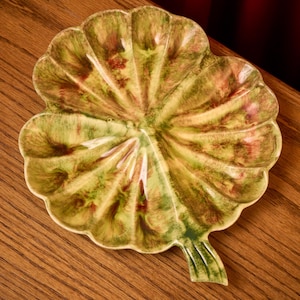 May include: A ceramic leaf-shaped serving dish with a scalloped edge. The dish features a blend of green, yellow, and reddish-brown hues, creating a natural, earthy aesthetic. The dish is placed on a wooden surface.