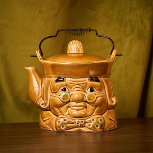 Może przedstawiać: Brązowy ceramiczny czajnik w kształcie ludzkiej głowy z czarnym metalowym uchwytem. Czajnik ma brązowy kapelusz, okulary i muszkę.