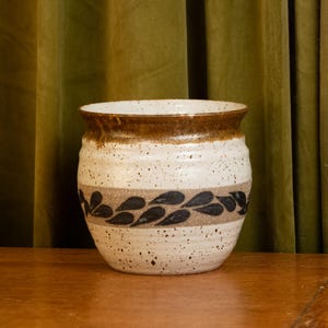 Könnte beinhalten: Eine gesprenkelte Keramikvase mit einer weiten Öffnung und einem braunen Rand. Die Vase hat ein Band aus braunen und schwarzen blattartigen Designs in der Mitte. Der Hintergrund ist ein grüner Vorhang.