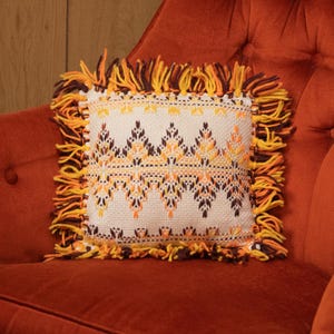 Può includere: Un cuscino quadrato con sfondo color crema e un motivo geometrico nei toni dell'arancione e del marrone. Il cuscino ha un bordo con frange gialle, arancioni e marroni. Il cuscino è appoggiato su una poltrona color ruggine.