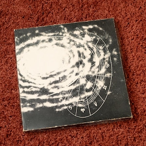 May include: A square, black box with a white galaxy design and a circular zodiac chart. The box is set against a reddish-brown, textured background. The design features astrological symbols and a swirling galaxy.