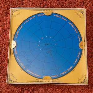 Può includere: Gioco da tavolo Astrometer con un design circolare blu con etichette delle case astrologiche e la scritta "Astrometer". Il tabellone è in una scatola color oro con disegni decorativi agli angoli. Lo sfondo è un tappeto rosso.