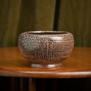 May include: A brown ceramic bowl with a textured surface and a decorative etched pattern of vertical lines. The bowl sits on a wooden table, showcasing its earthy tones and handcrafted design.