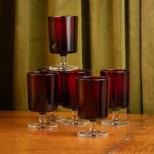 May include: A collection of six burgundy-colored wine glasses. Each glass has a short stem and a clear, circular base. The glasses are arranged on a wooden surface against a green curtain backdrop.