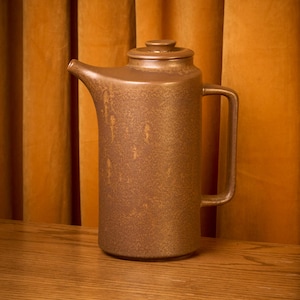 May include: A brown ceramic coffee pot with a lid, spout, and handle. The pot has a speckled texture and sits on a wooden surface against a golden-yellow curtain backdrop. The pot is approximately 12 inches tall.