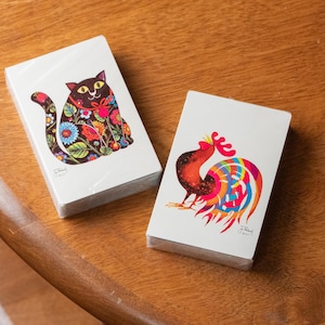 May include: Two decks of playing cards. One deck features a black cat with floral patterns, the other a colorful rooster. Both decks are on a wooden surface. The cards are wrapped in clear plastic.