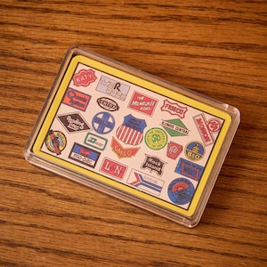 May include: A rectangular clear acrylic magnet featuring a yellow border and a collage of vintage railroad logos. Logos include "KATY", "THE ROCK", "Amtrak", and "Union Pacific Railroad". The magnet is displayed on a wooden surface.
