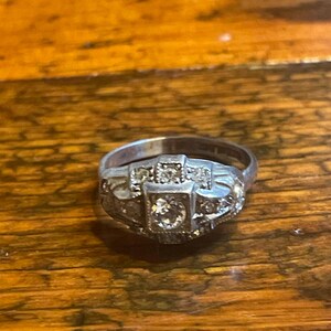 May include: A silver ring with a central square-cut diamond and smaller accent stones. The ring has an Art Deco design, with geometric shapes and intricate detailing. The ring is set on a wooden surface.