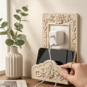 Pode incluir: Estação de carregamento de telefone montada na parede bege com detalhes em relevo floral. Um carregador branco está conectado à parede, com um telefone no suporte abaixo. Um pequeno vaso com vegetação à esquerda.