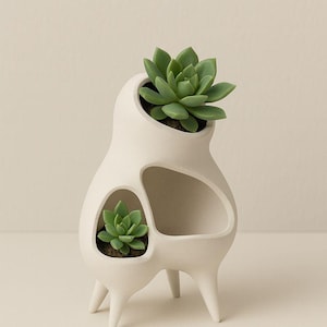 May include: A white ceramic planter with three leg supports, featuring three openings to hold plants. Two green succulents are planted in the openings. The planter has a modern, organic shape.