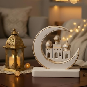 Peut inclure: Une décoration en forme de croissant de lune blanc avec des détails dorés et une conception de mosquée miniature. Une lanterne dorée avec une bougie allumée est à gauche. La scène est sur une table en bois sombre avec un éclairage doux.