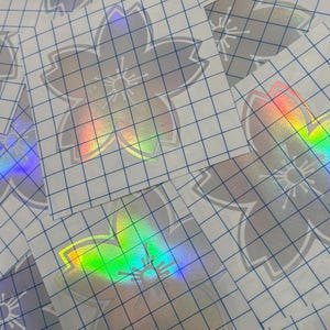 Könnte beinhalten: Holografische Blumenaufkleber mit Gitterrücken. Die Aufkleber zeigen ein Kirschblütendesign mit schillernden Regenbogenfarben. Die Aufkleber befinden sich auf einem weißen Hintergrund.