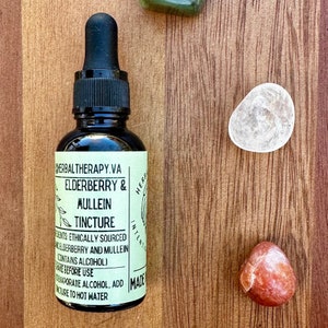 May include: A black glass bottle with a dropper, labeled "HERBALTHERAPY.VA ELDERBERRY & MULLEIN TINCTURE." The label is light green with black text. Two stones are next to the bottle on a wooden surface. The bottle contains a liquid.