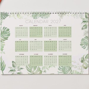 May include: A 2027 wall calendar featuring a white background with a border of green monstera and fern leaves. The calendar displays all twelve months of the year in a grid format with light green rectangular backgrounds for each month.