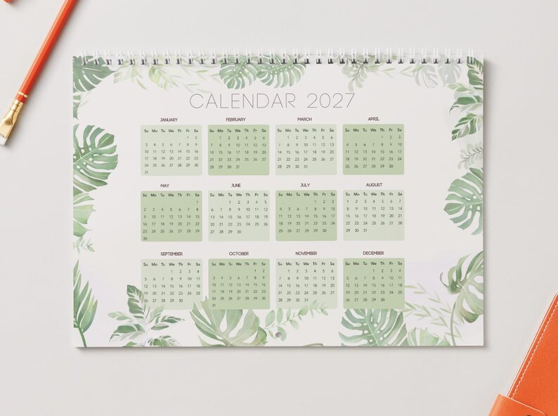 May include: A 2027 wall calendar featuring a white background with a border of green monstera and fern leaves. The calendar displays all twelve months of the year in a grid format with light green rectangular backgrounds for each month.