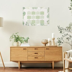 May include: A wall calendar for the year 2027 featuring a grid layout for twelve months. The calendar is decorated with a botanical pattern of green leaves and foliage surrounding the dates, printed on a white background.