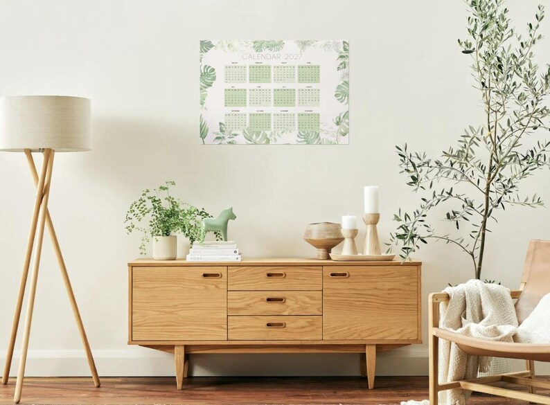 May include: A wall calendar for the year 2027 featuring a grid layout for twelve months. The calendar is decorated with a botanical pattern of green leaves and foliage surrounding the dates, printed on a white background.