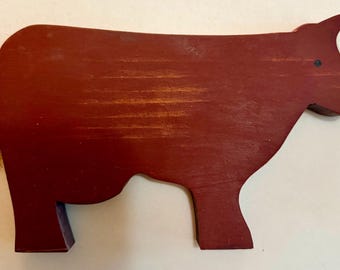 May include: A flat, cutout wooden cow figure painted in a deep, matte red finish. The silhouette features a simple, stylized body shape with a small black dot for an eye and a tuft of light brown, fibrous material attached to the rear as a tail.