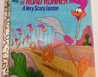 May include: A Little Golden Book titled The Road Runner A Very Scary Lesson featuring a large Road Runner character running on a desert road with three smaller Road Runners behind it, set against a blue sky and pink rock formations.