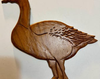 May include: A flat, brown, wooden cutout shaped like a goose in profile, facing left. The surface features carved lines detailing the feathers on the wing and the neck, with a smooth, polished finish throughout the entire body of the bird.