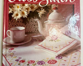 May include: A vintage book titled "Better Homes and Gardens: The Pleasures of Cross-Stitch." The cover features a still life with a teapot, teacup, and a cross-stitched tablecloth with floral patterns. The book is red with white text.