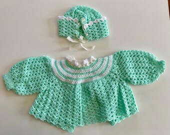 May include: A mint green crochet baby sweater and bonnet set. The sweater has a scalloped hem and sleeves, with white trim around the collar. The bonnet has a matching scalloped edge and white ribbon ties. The set is handmade.