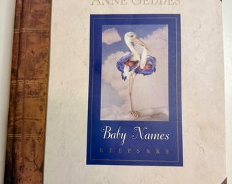May include: A hardcover book titled "Baby Names Keepsake" by Anne Geddes. The cover features a stork illustration within a blue frame against a cloudy sky. The book has a cream-colored cover with brown leather-like accents.