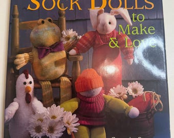 May include: A book cover titled "Adorable Sock Dolls to Make & Love" featuring handmade sock dolls. The dolls include a frog, a bunny, a chicken, and a doll wearing a hat. The cover also includes the authors' names, Connie Stone & Emola Lowe.