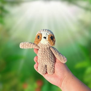 May include: A small crocheted sloth toy with brown fur, white chest, and black eyes. The sloth is being held in a person's hand.