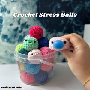 May include: A clear plastic bucket filled with colorful crochet stress balls. The balls are various colors, including pink, blue, green, and red. One pink ball with a blue center is held up. Text reads "Crochet Stress Balls" and "surprise or pick a color!"
