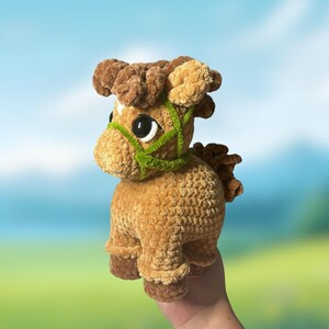 May include: A plush, crocheted horse toy in shades of brown and tan. The horse has a green halter and large black eyes. The toy is held up against a blurred background of a blue sky and green field.