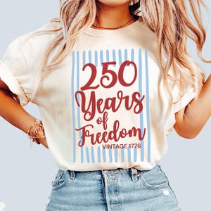 Woman wearing ivory Comfort Colors patriotic shirt featuring “250 Years of Freedom Vintage 1776” retro red and blue design for Fourth of July celebration