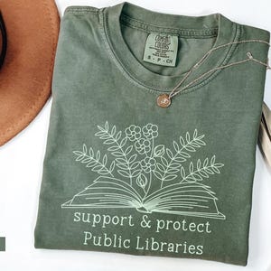 May include: A moss green t-shirt with a white outline of a book with flowers on top. The text "support & protect Public Libraries" is printed below the book.