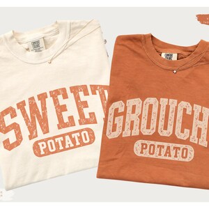 May include: Two short-sleeve t-shirts. The left shirt is ivory with orange distressed text reading SWEET POTATO. The right shirt is burnt orange with ivory distressed text reading GROUCHY POTATO. Both shirts feature a crew neckline and a Comfort Colors label.