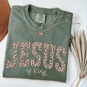 Op de afbeelding: Een mosgroen t-shirt met de tekst "JESUS is King" in een luipaardprint. Een gouden ketting en een gedroogde plant zijn ook zichtbaar. Het shirt is opgevouwen en heeft een Comfort Colors label.