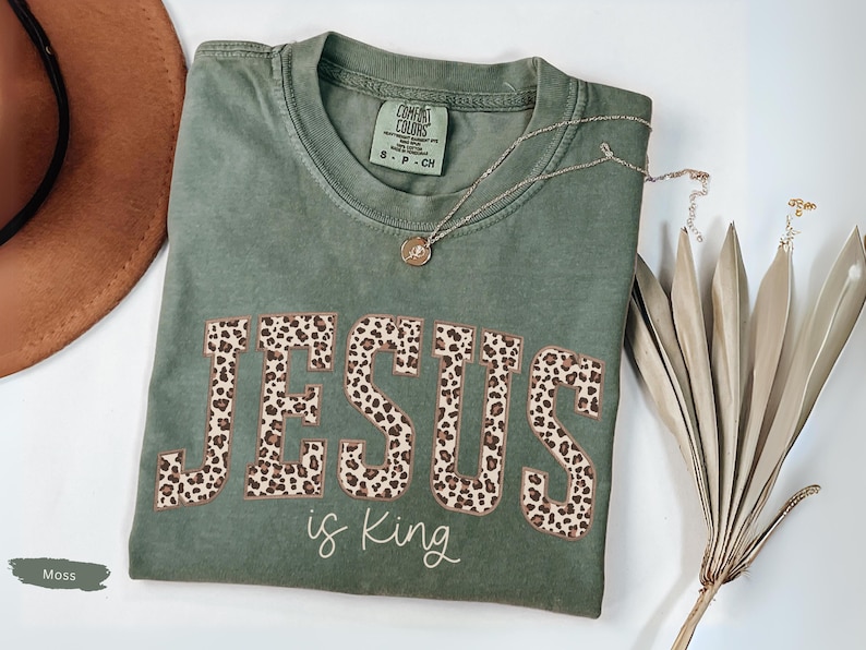 Op de afbeelding: Een mosgroen t-shirt met de tekst "JESUS is King" in een luipaardprint. Een gouden ketting en een gedroogde plant zijn ook zichtbaar. Het shirt is opgevouwen en heeft een Comfort Colors label.