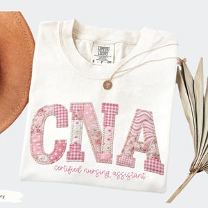 May include: Ivory t-shirt featuring the letters "CNA" in pink patchwork fabric. The text "Certified Nursing Assistant" is below the letters. A gold necklace and a brown hat are also visible.