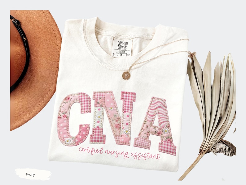 May include: Ivory t-shirt featuring the letters "CNA" in pink patchwork fabric. The text "Certified Nursing Assistant" is below the letters. A gold necklace and a brown hat are also visible.