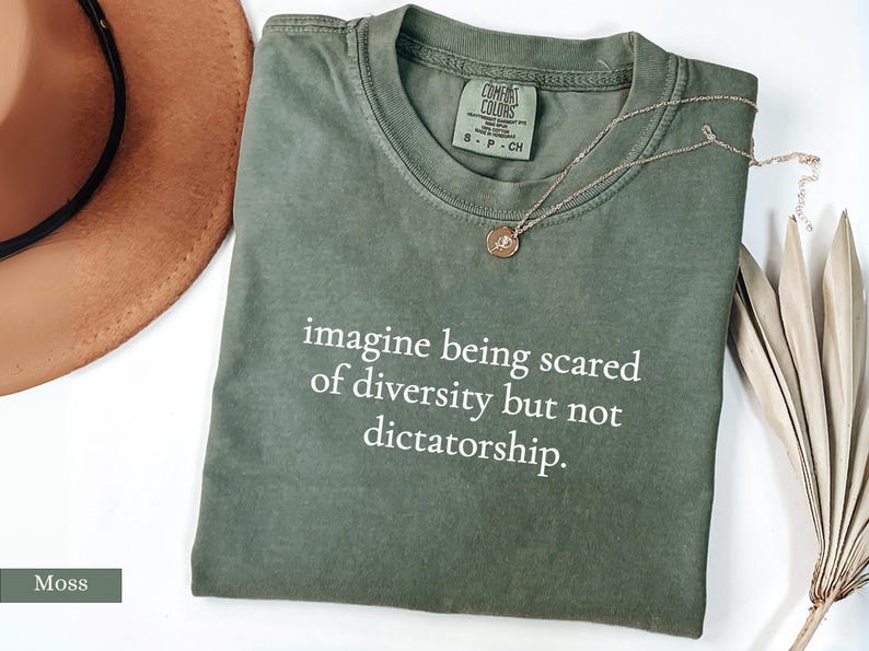 May include: A moss green t-shirt with the text "imagine being scared of diversity but not dictatorship." A gold necklace and a brown hat are also visible. The shirt has a Comfort Colors tag.