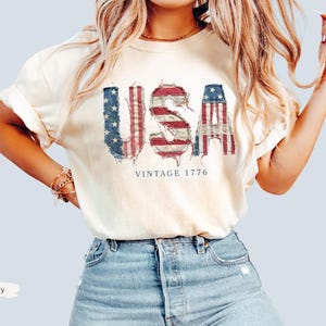 Puede incluir: Camiseta color marfil con la palabra "USA" en un dise&ntilde;o desgastado y patri&oacute;tico con colores rojo, blanco y azul. El texto "VINTAGE 1776" est&aacute; impreso debajo. La camiseta se combina con pantalones cortos vaqueros azules.