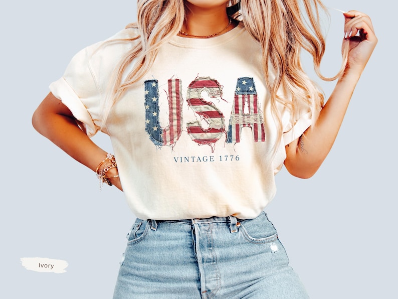 Puede incluir: Camiseta color marfil con la palabra "USA" en un dise&ntilde;o desgastado y patri&oacute;tico con colores rojo, blanco y azul. El texto "VINTAGE 1776" est&aacute; impreso debajo. La camiseta se combina con pantalones cortos vaqueros azules.