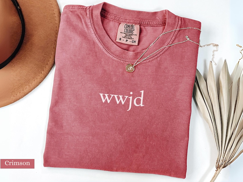 Puede incluir: Camiseta de color carmes&iacute; con el texto blanco "wwjd". Un collar plateado con un colgante descansa sobre la camisa. La camisa tiene una etiqueta que dice "Comfort Colors". Un sombrero marr&oacute;n y una planta seca est&aacute;n en el fondo.