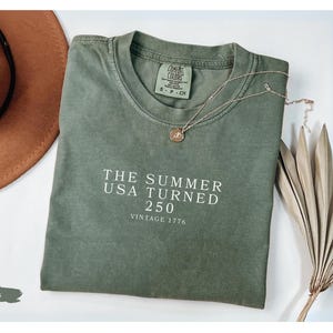 May include: A moss green t-shirt with the text "THE SUMMER USA TURNED 250 VINTAGE 1776" in white. A brown hat and a gold necklace with a pendant are next to the shirt. Dried decorative plants are also in the image.