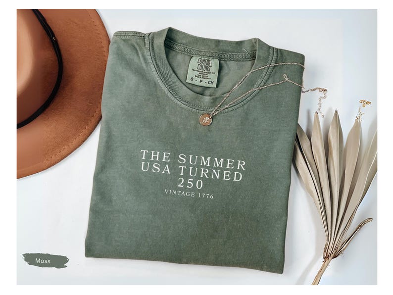 May include: A moss green t-shirt with the text "THE SUMMER USA TURNED 250 VINTAGE 1776" in white. A brown hat and a gold necklace with a pendant are next to the shirt. Dried decorative plants are also in the image.