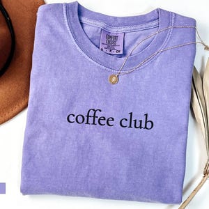 May include: A lilac-coloured t-shirt with the words "coffee club" printed in black. A gold necklace with a pendant is draped over the shirt. A brown hat and dried palm leaves are in the background.