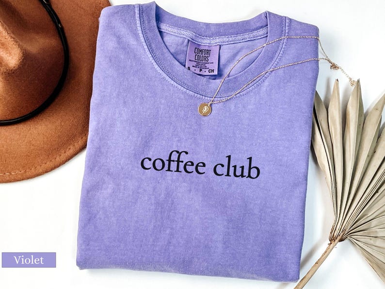 May include: A lilac-coloured t-shirt with the words "coffee club" printed in black. A gold necklace with a pendant is draped over the shirt. A brown hat and dried palm leaves are in the background.