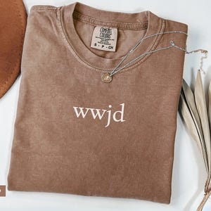 Puede incluir: Camiseta color espresso doblada con el texto "wwjd" en blanco. Tiene una etiqueta Comfort Colors. Un collar plateado con un peque&ntilde;o colgante y plantas secas decorativas est&aacute;n al lado de la camiseta.