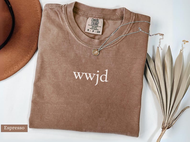 Puede incluir: Camiseta color espresso doblada con el texto "wwjd" en blanco. Tiene una etiqueta Comfort Colors. Un collar plateado con un peque&ntilde;o colgante y plantas secas decorativas est&aacute;n al lado de la camiseta.