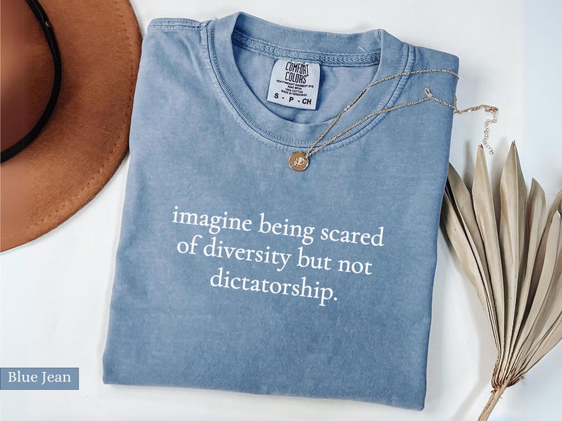 May include: A blue jean colored t-shirt with the text "imagine being scared of diversity but not dictatorship." A gold necklace with a pendant rests on the shirt. A brown hat and dried plant are in the background.